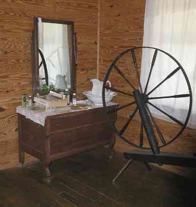 Cantonment:-Roy-Hyatt-Environmental-Center-Dog-Trot-House_12.jpg:  iron bed, quilt, ragdoll, bedroom, wooden walls, wood floor, spinning wheel, dresser, rocking chair