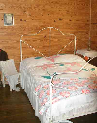 Cantonment:-Roy-Hyatt-Environmental-Center-Dog-Trot-House_13.jpg:  iron bedstread, oil lamp, iron bed, quilt, ragdoll, bedroom, wooden walls, wood floor, rocking chair